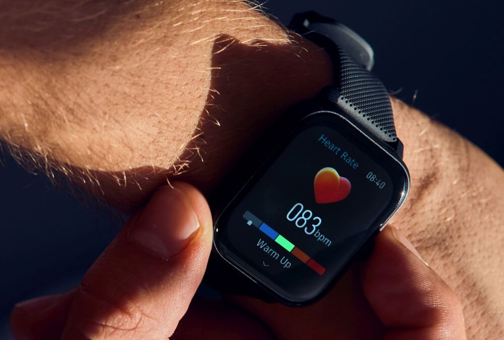 Na zdjęciu widać smartwatch na nadgarstku, wyświetlający pomiar tętna 083 bpm z ikoną serca. Ekran pokazuje również napis \'Heart Rate\' oraz \'Warm Up\'.