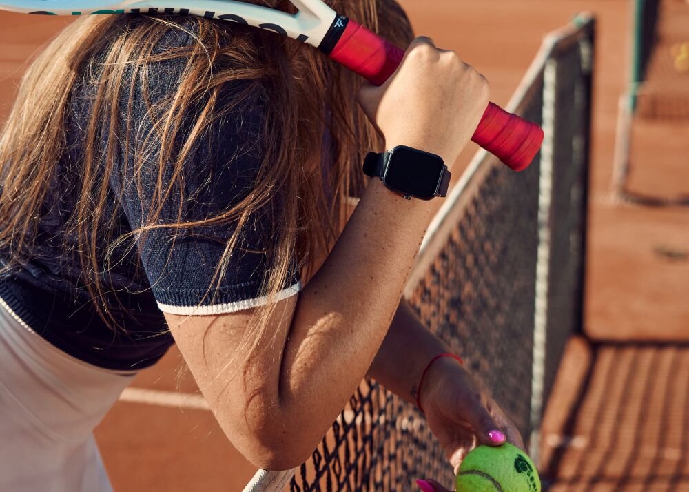 Osoba trzyma rakietę tenisową i piłkę, na nadgarstku ma czarny smartwatch. W tle widoczne jest ogrodzenie kortu tenisowego.