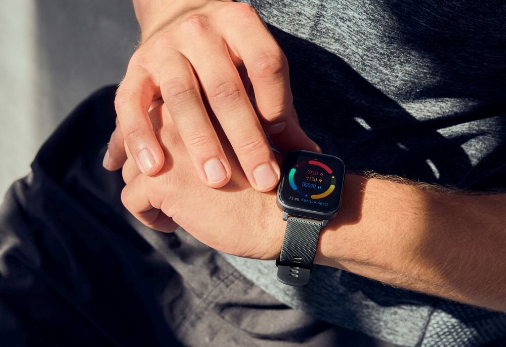 Osoba nosi czarny smartwatch z kolorowym wyświetlaczem na nadgarstku. Na ekranie widoczne są kolorowe wskaźniki i tekst: \'100% 0 DZI 0 KROK\'.