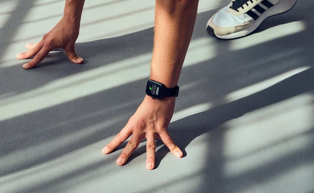 Osoba w pozycji do pompek nosi czarny smartwatch na nadgarstku. W tle widać cień kratownicy na podłodze.