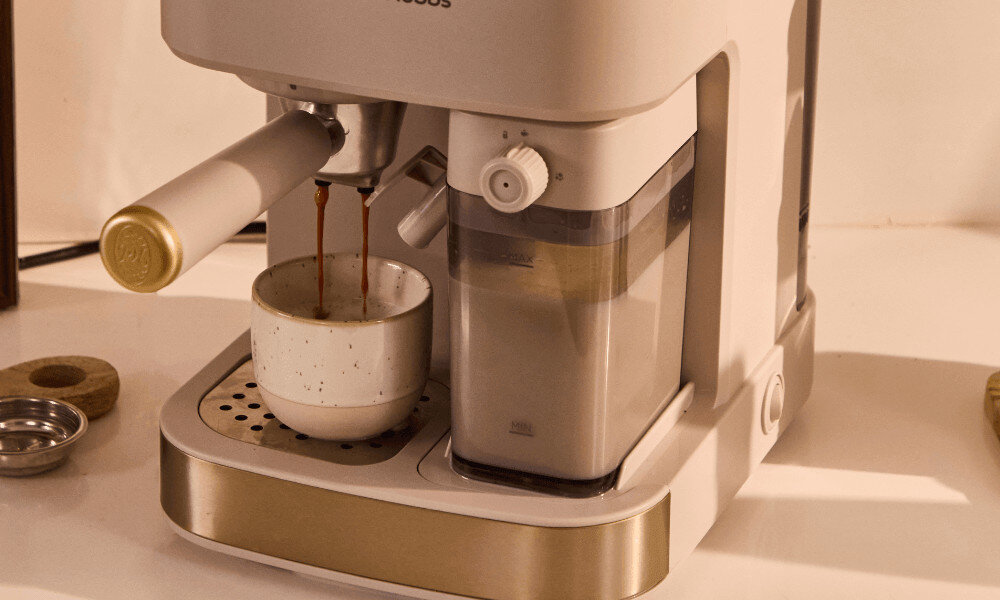 Kremowy ekspres do kawy z logo \'Russell Hobbs\' przygotowuje kawę, która spływa do białej filiżanki. Obok znajduje się przezroczysty pojemnik na wodę z miarką.