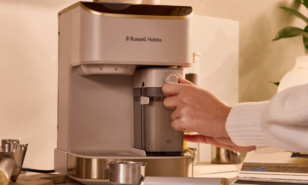 Kremowy ekspres do kawy marki Russell Hobbs z widocznym logo, użytkowany przez osobę. W tle znajdują się elementy kuchenne i rośliny.