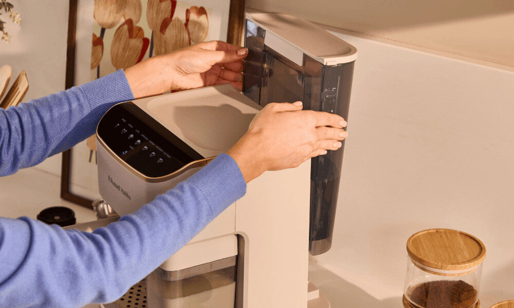 Osoba w niebieskim swetrze obsługuje kremowy ekspres do kawy z widocznym logo \'Russell Hobbs\'. Obok ekspresu stoi szklany pojemnik z drewnianą pokrywką.