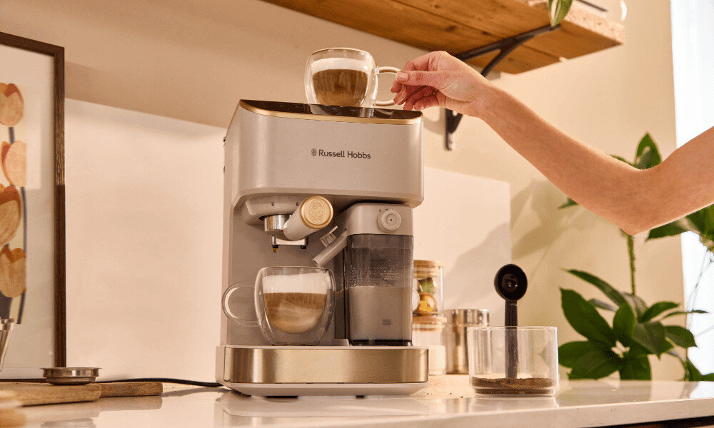 Ekspres do kawy w kolorze kremowym z logo \'Russell Hobbs\', na którym przygotowywana jest kawa w szklanej filiżance. W tle widoczne są elementy kuchenne, takie jak rośliny i półka.