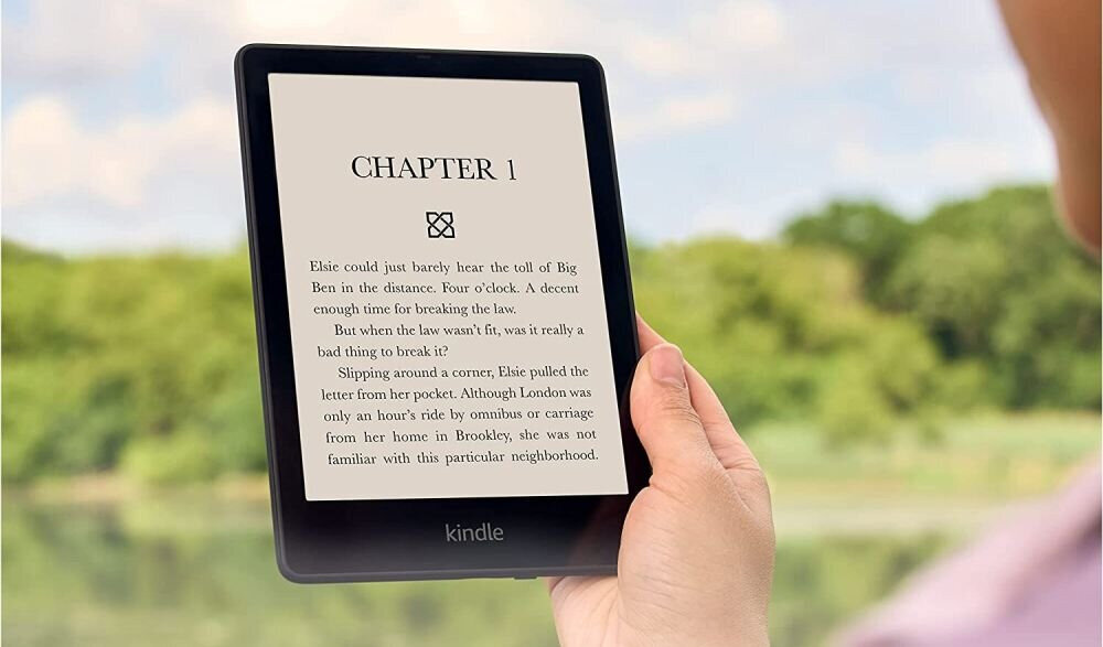 Osoba trzyma czytnik e-booków z widocznym tekstem na ekranie: \'CHAPTER 1\'. W tle znajduje się rozmyty krajobraz z zielenią.