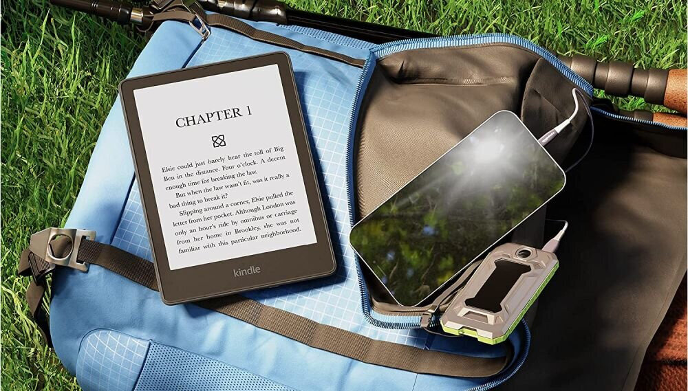 Na obrazie widoczny jest czytnik e-booków z wyświetlonym tekstem \'CHAPTER 1\'. Obok znajduje się smartfon i powerbank, wszystko umieszczone na niebieskim plecaku na trawie.