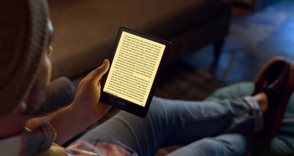 Osoba trzyma czytnik e-booków z podświetlonym ekranem, na którym widoczny jest tekst. Czytnik ma czarną obudowę i logo \'Kindle\' na dole ekranu.
