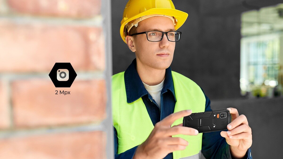 Osoba w kasku i kamizelce trzyma czarny telefon. Obok widoczna jest ikona aparatu z napisem \'2 Mpx\'.