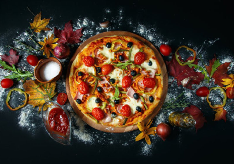 Na ciemnym tle znajduje się pizza z pomidorami, oliwkami i serem, otoczona mąką, liśćmi i warzywami. Obok pizzy widoczny jest sos pomidorowy w szklanym naczyniu oraz mała miseczka z solą.