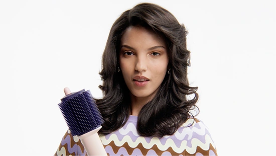 Młoda, uśmiechnięta modelka trzyma w dłoni szczotko-suszarkę Dyson Airsmooth i prezentuje gotową, wystylizowaną fryzurę.