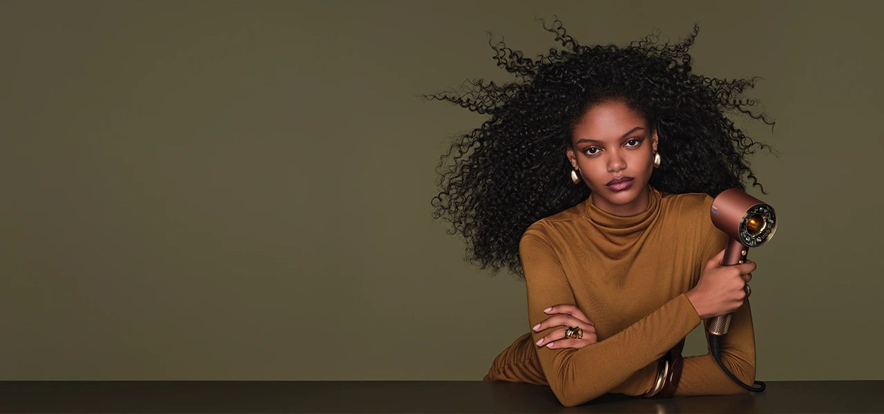 Modelka o długich, gęstych włosach w stylu afro trzyma w ręku suszarkę Dyson Supersonic Nural skierowaną na wprost włosów