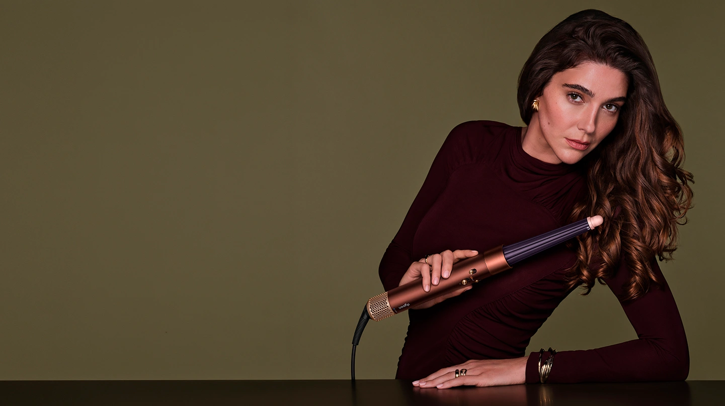 Modelka z długimi i ułożonymi w fale włosami trzyma w ręku multistyler Dyson Airwrap i.d. w kolorze miedzianym.