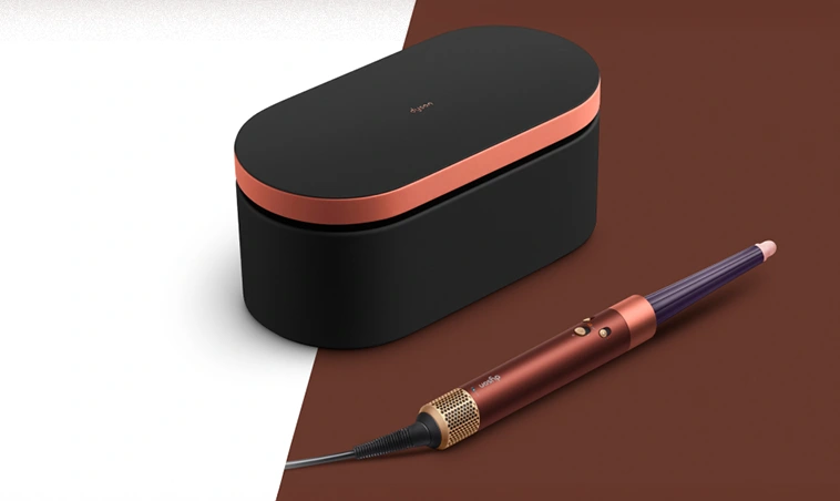 Etui oraz miedziany multistyler Dyson Airwrap ze stożkową końcówką leżą na beżowo-brązowym blacie.