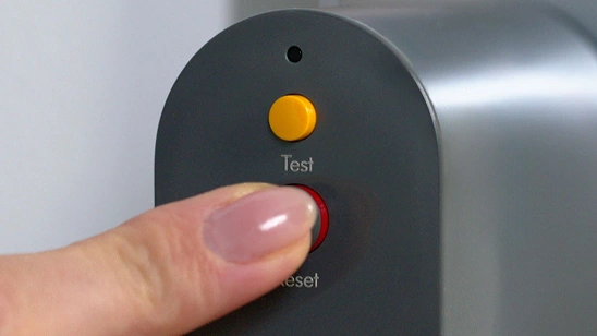Palec naciska czerwony przycisk oznaczony jako \'Reset\' na szarej powierzchni. Nad nim znajduje się żółty przycisk z napisem \'Test\'.