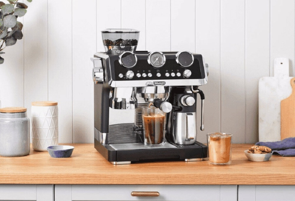 Na drewnianym blacie stoi ekspres do kawy marki DeLonghi z filiżanką kawy i metalowym dzbankiem. Obok znajdują się pojemniki, filiżanka i talerzyk z ciastkami.