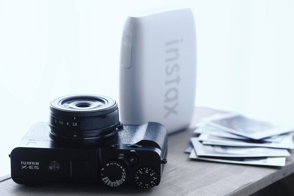 Na drewnianym stole znajduje się czarny aparat bezlusterkowy z widocznym obiektywem i pokrętłami. Obok aparatu stoi biały drukarka z napisem \'instax\' oraz kilka zdjęć.