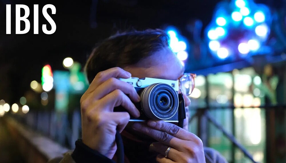 Osoba trzyma aparat fotograficzny, a w tle widać rozmyte światła. Na obrazie widoczny jest tekst \'IBIS\'.