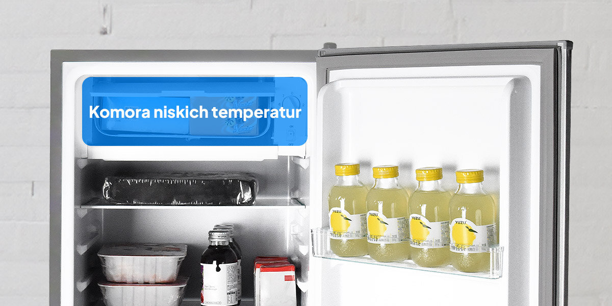 Otwarta lodówka z widocznymi produktami spożywczymi, w tym butelkami napoju na drzwiach. Na górnej półce znajduje się napis \'Komora niskich temperatur\'.