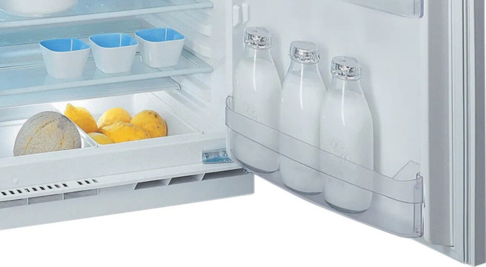 Wnętrze lodówki z trzema półkami, na których znajdują się niebieskie pojemniki, cytryny i melon. Na drzwiach lodówki umieszczone są trzy butelki z mlekiem.