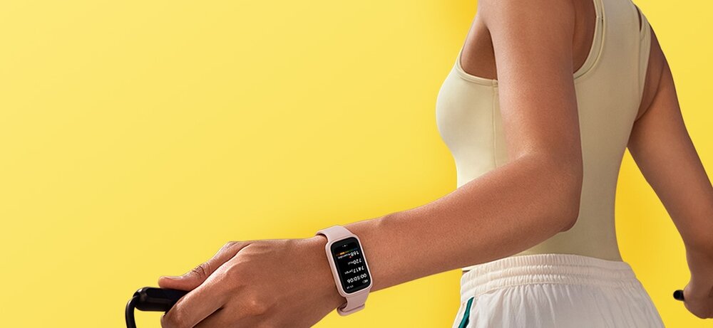 Osoba w jasnym stroju sportowym nosi beżowo-biały smartband na nadgarstku. Tło jest jednolite, żółte.