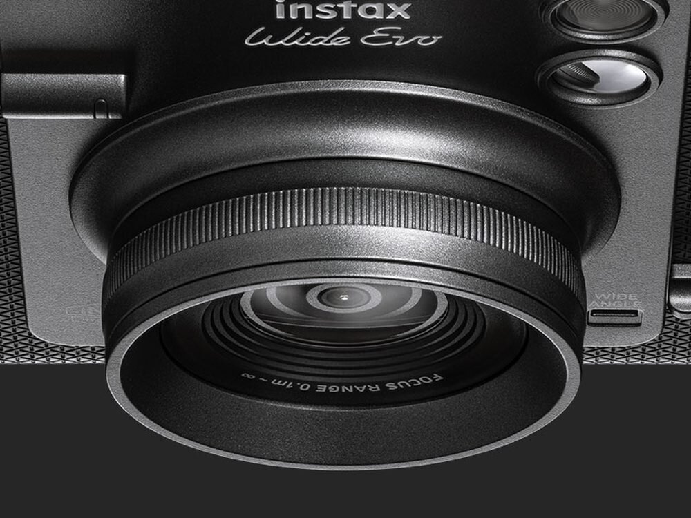 Zbliżenie na obiektyw aparatu z widocznym napisem \'instax Wide Evo\'. Na obiektywie znajduje się tekst \'FOCUS RANGE 0.1m - ∞\'.