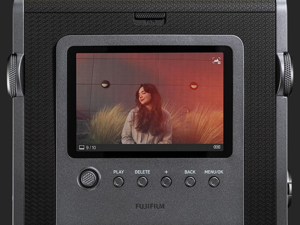 Tył aparatu z wyświetlaczem pokazującym zdjęcie osoby na tle roślin. Widoczne przyciski: PLAY, DELETE, +, BACK, MENU/OK oraz logo FUJIFILM.