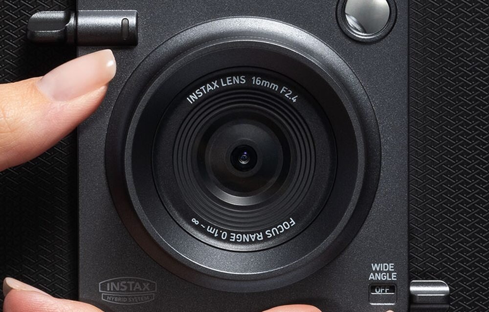 Zbliżenie na obiektyw aparatu z napisem \'INSTAX LENS 16mm F2.4 FOCUS RANGE 0.1m - ∞\'. Widać także logo \'INSTAX\' oraz napis \'WIDE ANGLE\'.