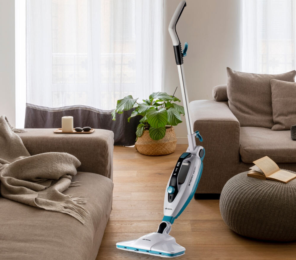 W jasnym salonie z drewnianą podłogą stoi biały mop parowy z niebieskimi akcentami. W tle znajdują się beżowe kanapy, roślina w koszu i zasłonięte okno.