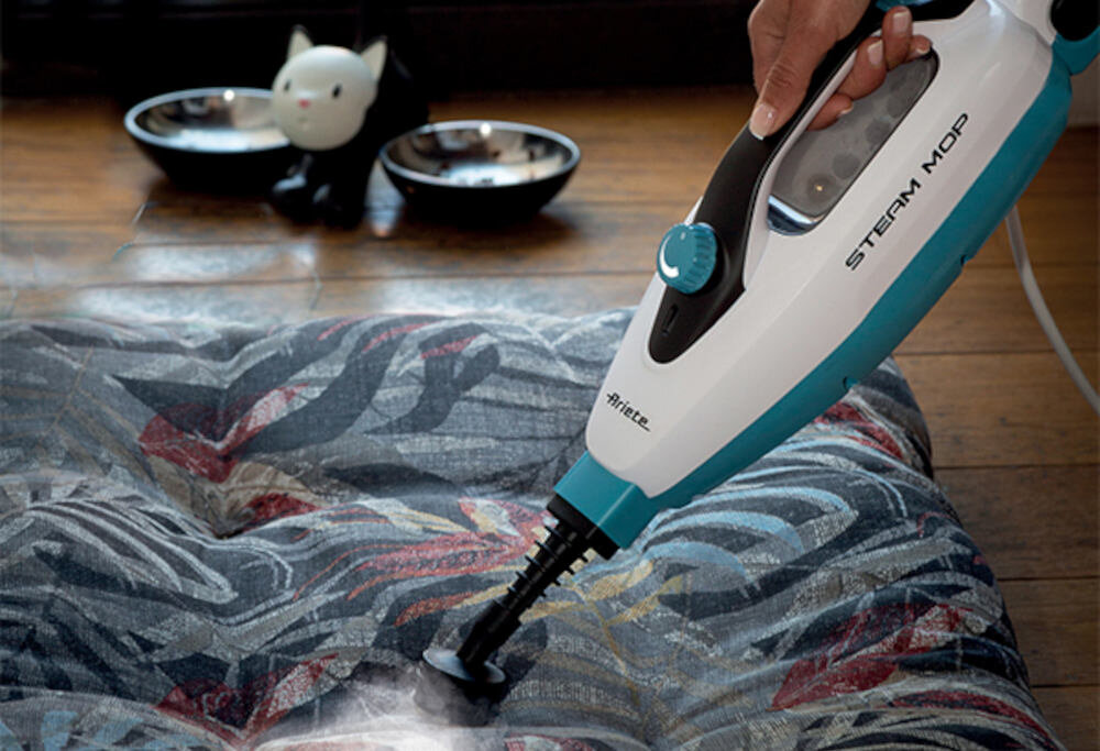 Osoba używa białego mopa parowego z niebieskimi elementami na kolorowej tkaninie. Na mopie widoczne są napisy \'STEAM MOP\' i \'ARIETE\'.
