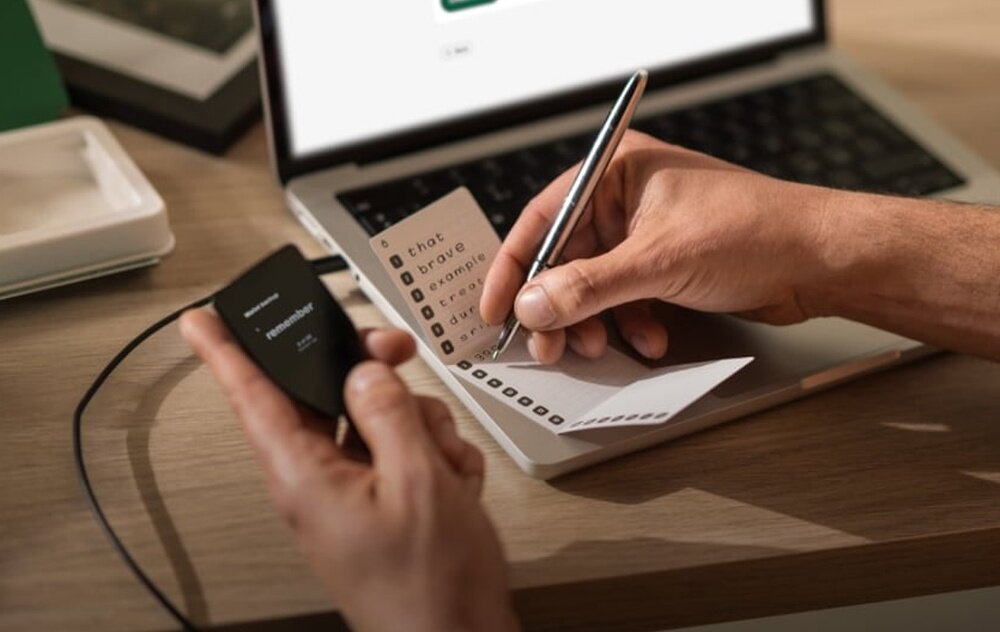 Osoba trzyma urządzenie z napisem \'TREZOR\' i \'trezor.io/start\', jednocześnie zapisując słowa na kartce papieru. W tle znajduje się otwarty laptop.