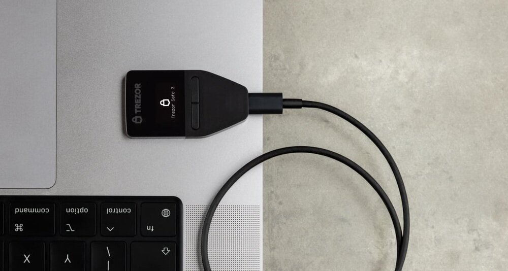 Na obrazie widoczny jest czarny portfel kryptowalut TREZOR Safe 3 podłączony do laptopa za pomocą kabla USB. Na ekranie urządzenia wyświetlona jest ikona kłódki i napis \'TREZOR\'.