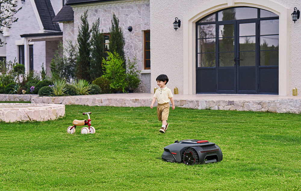 Na trawniku przed domem znajduje się robot koszący oraz dziecko obok rowerka. W tle widoczna jest fasada budynku z dużymi oknami i roślinnością.
