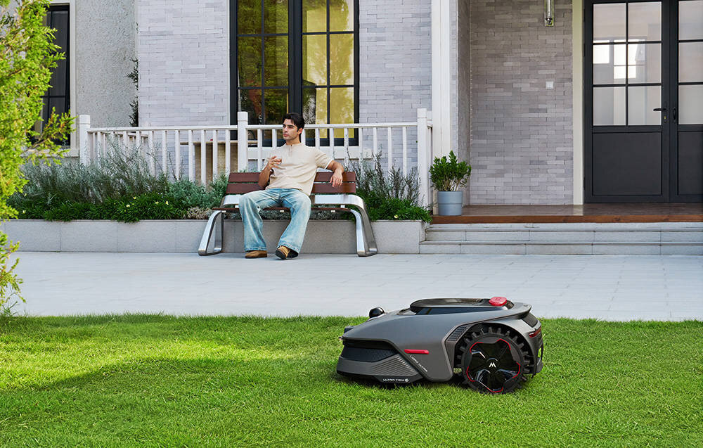 Na trawniku znajduje się robot koszący o nowoczesnym designie. W tle widać osobę siedzącą na ławce przed domem.