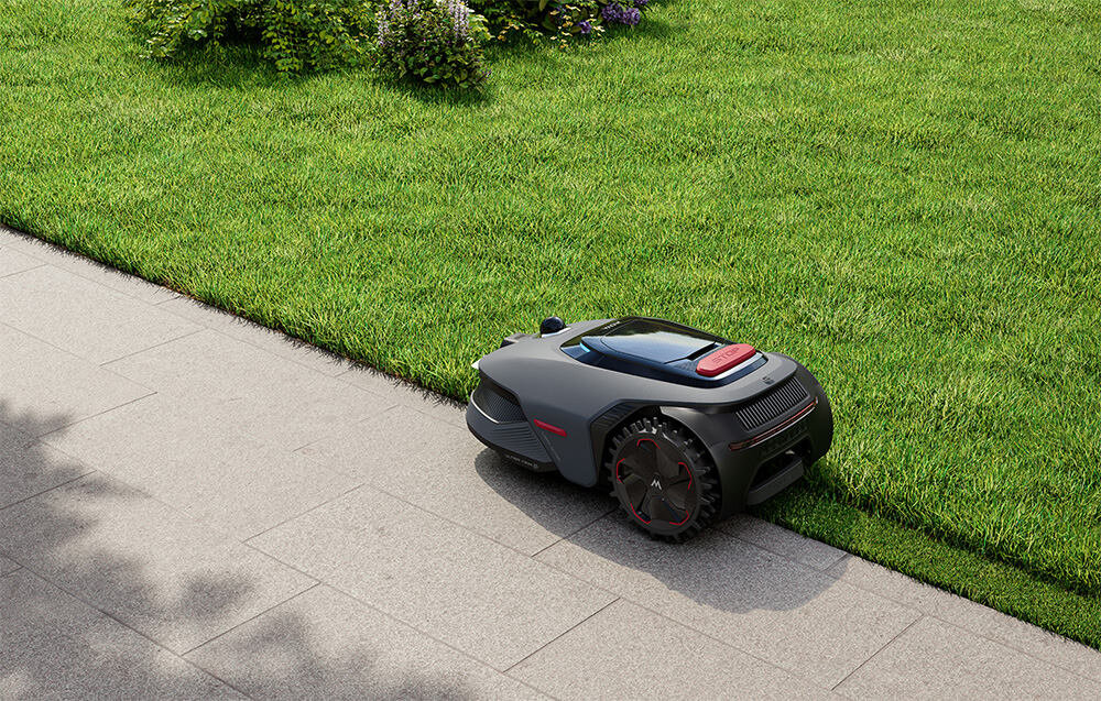 Robot koszący porusza się po krawędzi trawnika obok chodnika. Urządzenie ma nowoczesny design z ciemną obudową i czerwonymi akcentami.