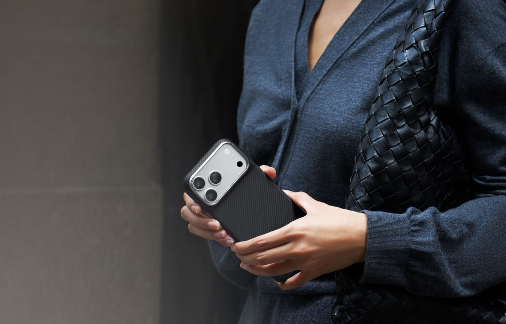 Osoba trzyma smartfon w czarno-szarym etui z trzema widocznymi obiektywami aparatu. Ubrana jest w ciemną odzież i trzyma plecioną torbę.