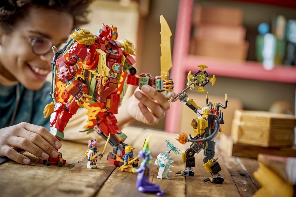 Na drewnianym stole znajdują się figurki LEGO, w tym duży czerwono-złoty mech oraz kilka mniejszych postaci. W tle widać osobę uśmiechającą się i bawiącą się zestawem.