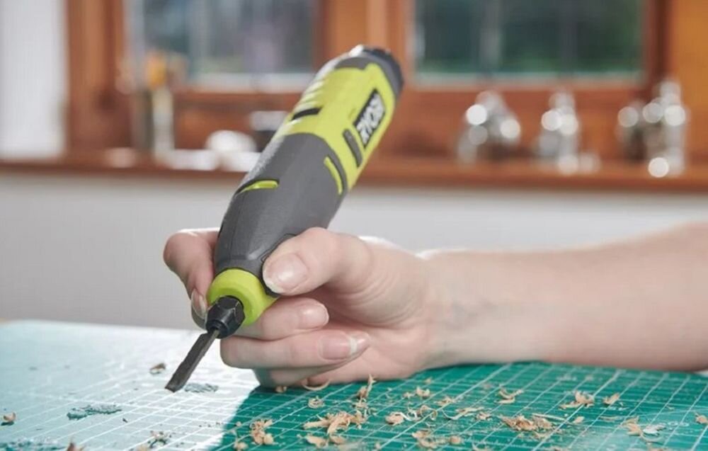 Osoba trzyma grawerkę w kolorze zielono-szarym z widocznym logo \'RYOBI\'. Na stole znajdują się wióry i zielona mata do cięcia.
