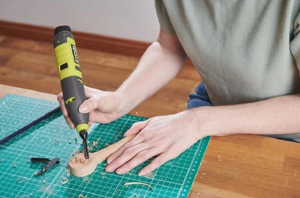 Osoba używa grawerki do pracy nad drewnianym przedmiotem na zielonej macie do cięcia. Na grawerce widoczne jest logo \'RYOBI\'.