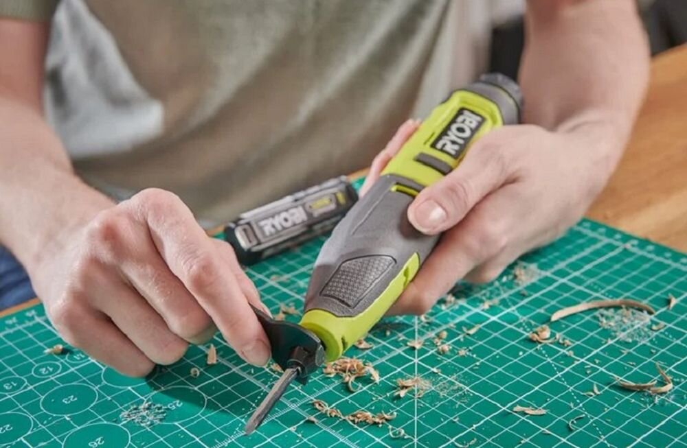 Osoba trzyma grawerkę z widocznym logo \'RYOBI\' i pracuje na zielonej macie do cięcia, wokół której znajdują się wióry. Obok leży bateria z napisem \'RYOBI\'.