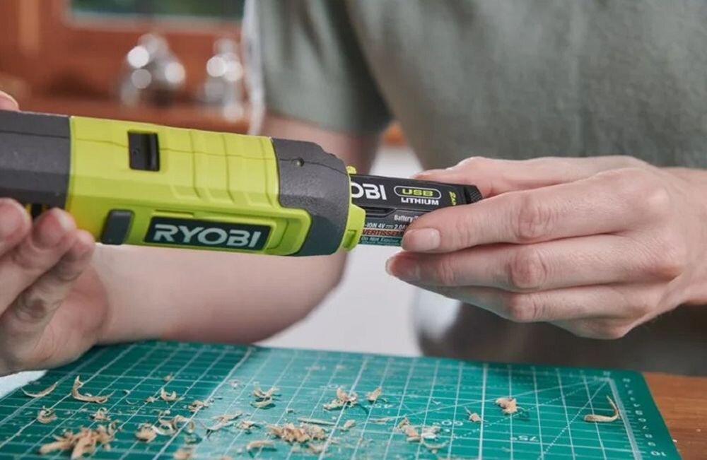 Osoba trzyma grawerkę z widocznym logo \'RYOBI\' oraz baterię oznaczoną jako \'USB Lithium\'. Na stole leży zielona mata z resztkami drewna.