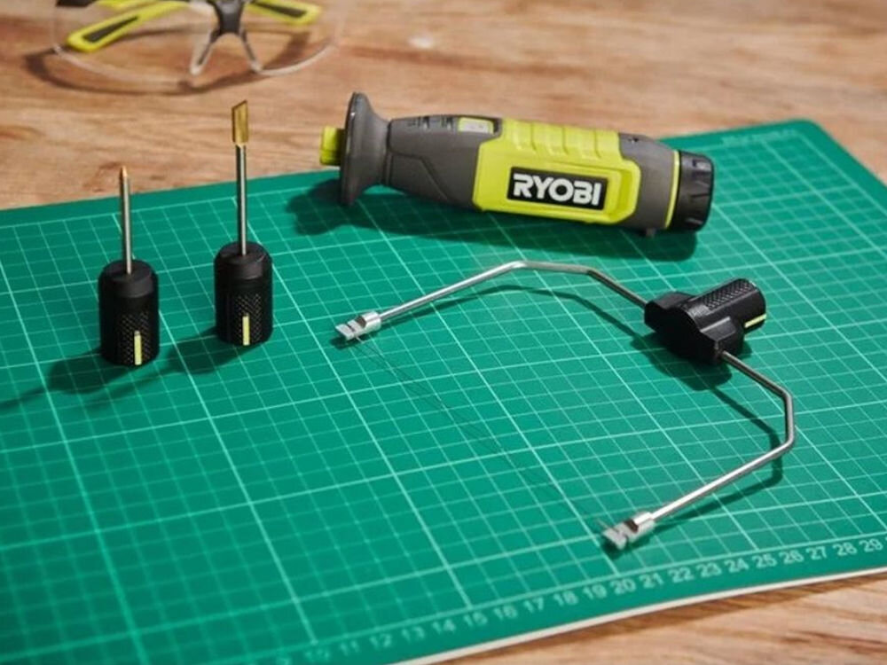 Na zielonej macie do cięcia leży narzędzie z logo \'RYOBI\' oraz dwa wymienne ostrza i drut tnący. Narzędzie ma szaro-żółtą obudowę.