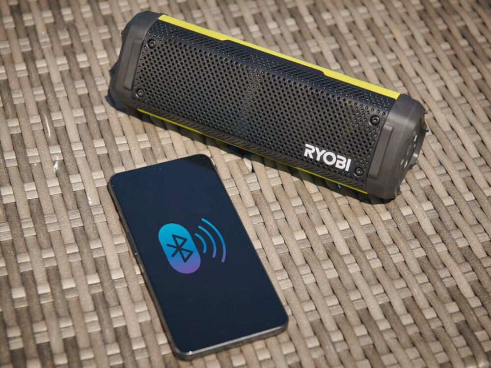Na plecionym tle znajduje się głośnik mobilny z logo \'RYOBI\' oraz smartfon z ikoną Bluetooth na ekranie.