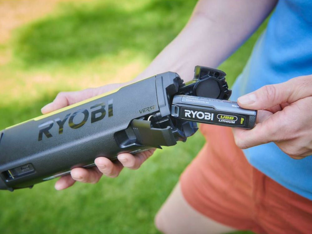 Osoba trzyma głośnik mobilny marki RYOBI, do którego wkłada baterię oznaczoną jako \'RYOBI USB Lithium\'.