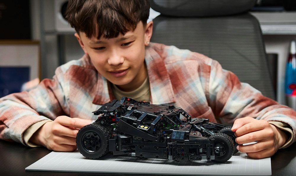 Osoba siedzi przy stole i trzyma model pojazdu z klocków LEGO, przypominający Batmobile Tumbler. Model jest czarny i szczegółowo wykonany.