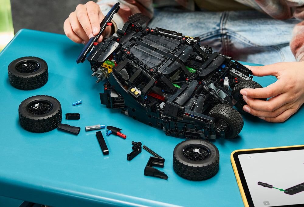 Osoba składa model samochodu z klocków LEGO na niebieskim stole, wokół leżą części i koła. Obok znajduje się tablet z instrukcją montażu.