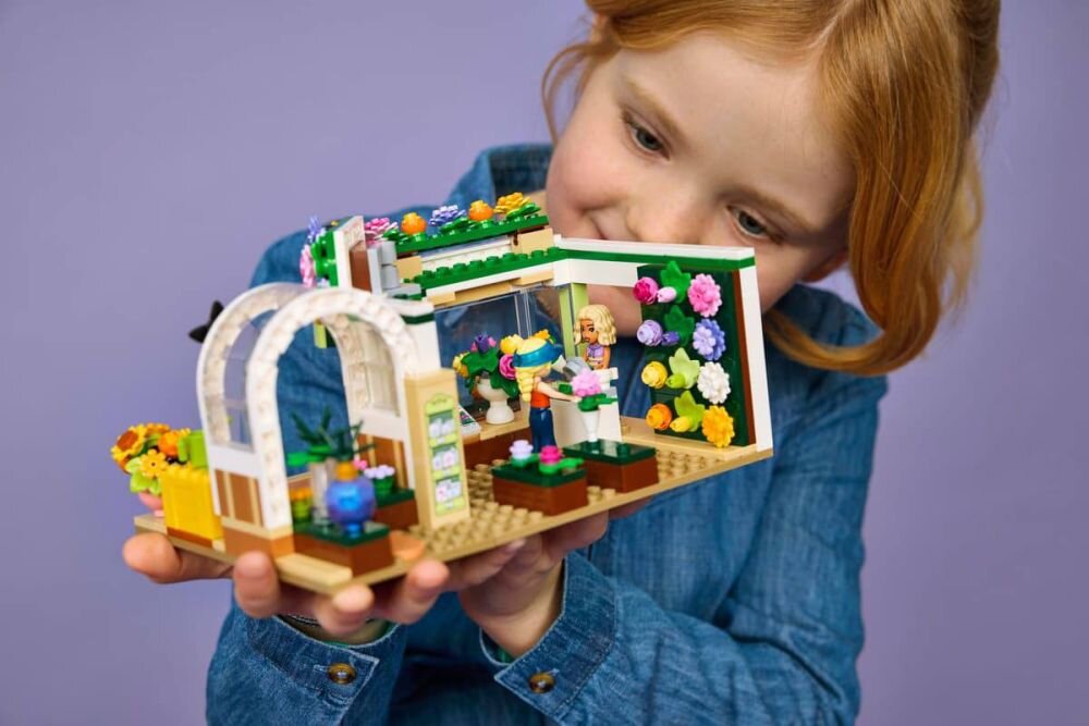 Dziecko trzyma zestaw LEGO przedstawiający kwiaciarnię z kolorowymi kwiatami i figurkami. Tło jest jednolite, fioletowe.