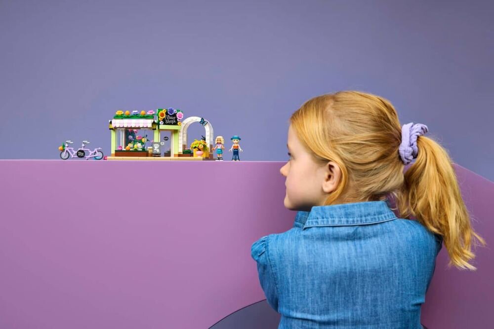 Dziewczynka w niebieskiej koszuli patrzy na zestaw LEGO przedstawiający kwiaciarnię z figurkami i rowerem na fioletowym tle.