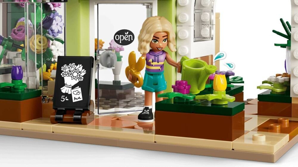 Zestaw LEGO przedstawia kreatywną kwiaciarnię z figurką postaci trzymającą konewkę i kwiaty. Na drzwiach widoczny jest napis \'open\', a obok stoi tablica z rysunkiem bukietu i oznaczeniem \'5+\'.