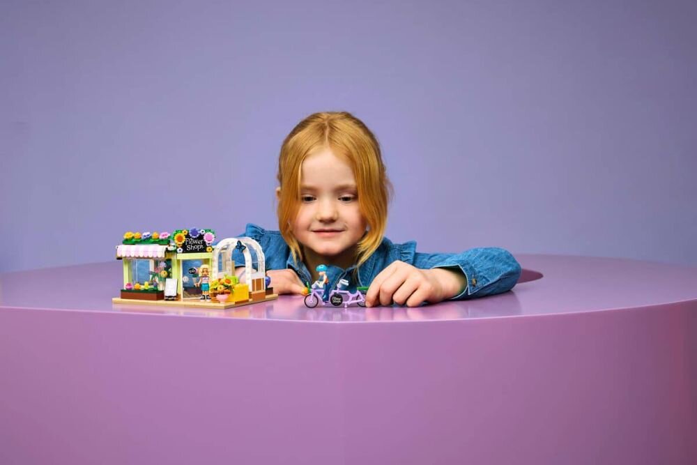 Dziecko bawi się zestawem LEGO przedstawiającym kwiaciarnię i figurkę na rowerze na fioletowym stole. W tle znajduje się jednolite, fioletowe tło.