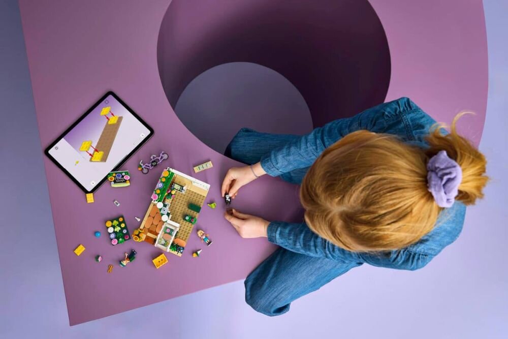 Osoba siedzi przy dużym, fioletowym stole, układając zestaw klocków LEGO. Obok znajduje się tablet z wyświetlonym obrazem klocków.